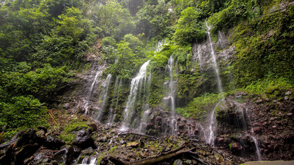 Misty multi-tier waterfall flowing over moss-covered rocks in a lush tropical forest, creating a peaceful natural landscape with rich greenery. © Aji Gallery