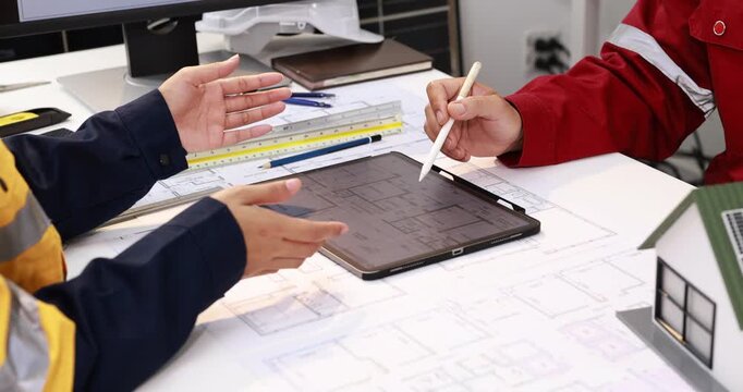 Construction engineers review building plans on a tablet at a desk, discussing measurements and technical drawings in an office.