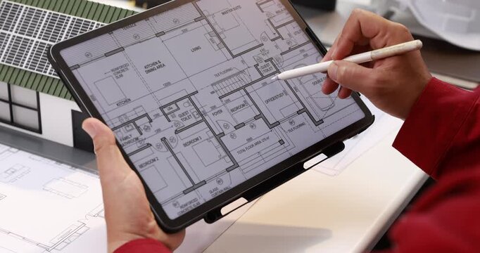 Close view of an architect holding a tablet with a digital house floor plan and pointing at the layout with a stylus at a design desk.