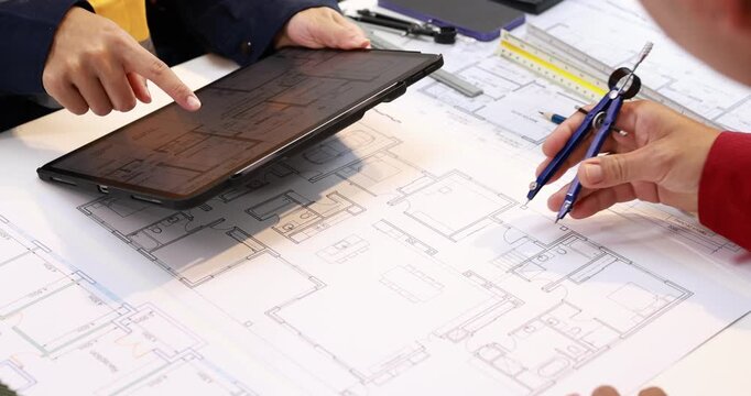 Architects review detailed building floor plans at a desk, using a tablet, ruler, compass and pen for project planning.