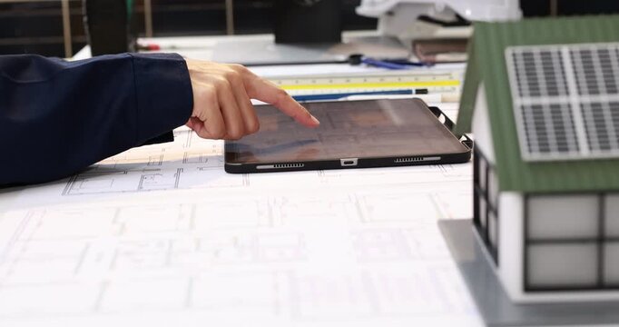 Close-up of an architect using a tablet on building plans at a desk, with document folders in the foreground.