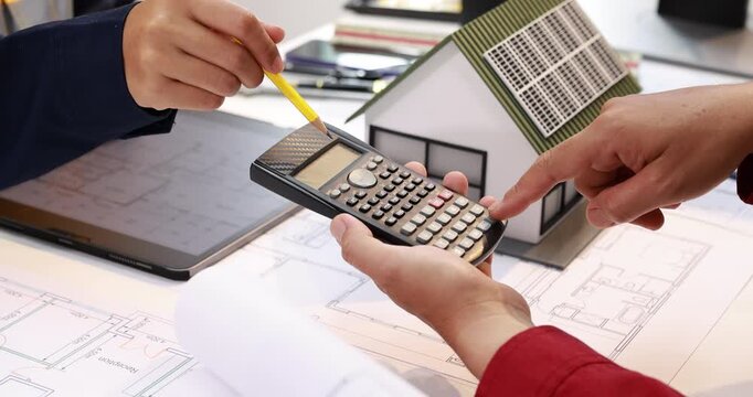 Architects review house plans with a calculator, model home and documents on a desk, planning construction costs and a residential building project.