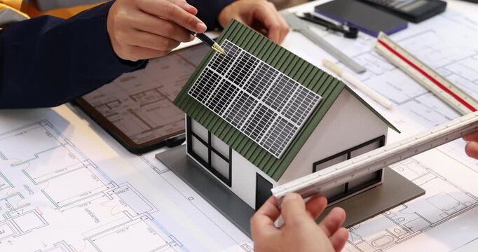 Architects examine a small house model with rooftop solar panels over building plans, measuring the design for an energy-efficient home.