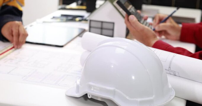 White safety helmet on architectural plans with rolled blueprints and professionals discussing a house model at a construction planning meeting.