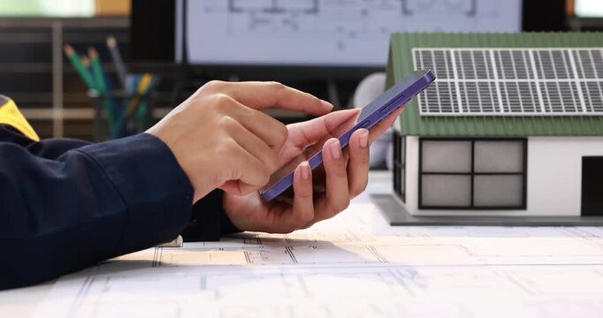 Architect using a tablet beside a model house with solar panels and building plans, sustainable home design and energy planning.