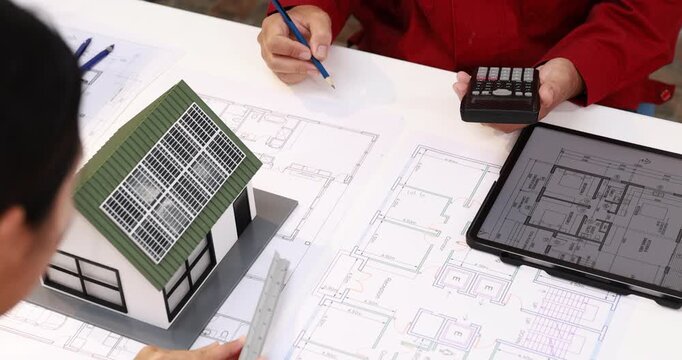 Architects review house plans with a model home, calculator and tablet during a building design meeting.