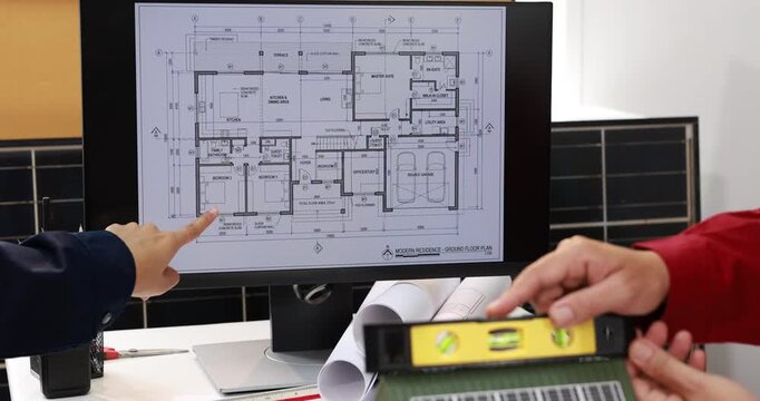 Architects review a house floor plan on a computer monitor while measuring printed drawings with a spirit level in a modern office.