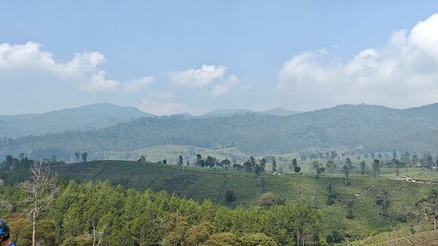 Panoramic green hills and agricultural fields stretch toward distant mountains beneath a bright blue sky with scattered clouds, showcasing a peaceful natural landscape.