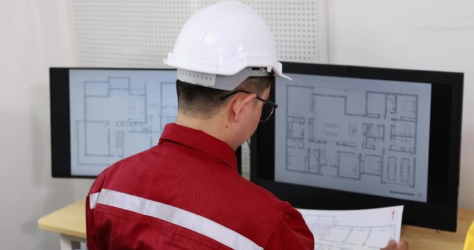 Rear view of a construction engineer in a hard hat reviewing building plans on dual computer monitors in an office.