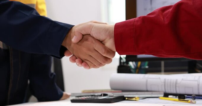 Close-up of two business people shaking hands across an office desk with building plans, calculator and pens, a successful agreement.