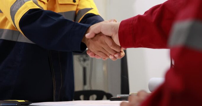 Close-up of workers in protective uniforms shaking hands across a desk, agreement, teamwork and professional cooperation in an industrial workplace.