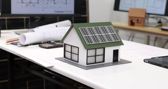 Small house model with rooftop solar panels on an architect desk, surrounded by building plans, drawings and a computer screen.