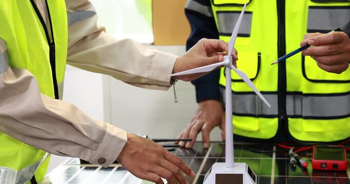 Engineers in safety vests review a small wind turbine model and solar panel during a renewable energy project meeting.