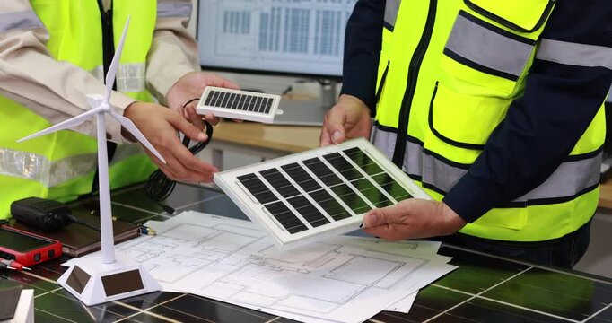 Engineers in safety vests review solar panels, a wind turbine model and building plans for a renewable energy project.