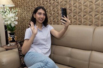 Portrait of a attractive young indian girl taking selfie picture while sitting on cozy sofa in living room at home. Say Hi. © Pix4Ads