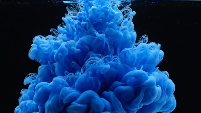 Blue ink drops falling into water and blooming into swirling clouds on a black background