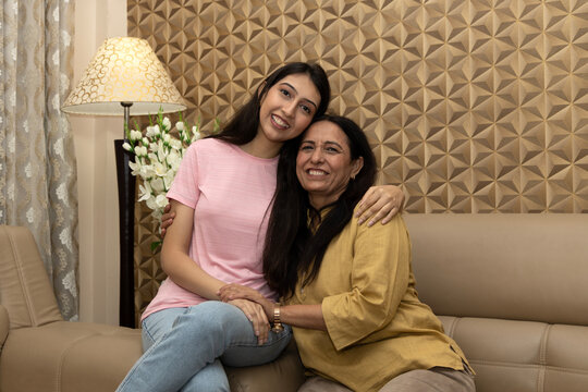 Happy Beautiful daughter hugging mother at home, portrait. Smiling mother and daughter bonding together looking at camera sitting on sofa.