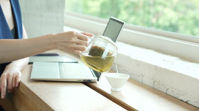 Woman pouring herbal tea from a glass teapot beside a laptop on a brig