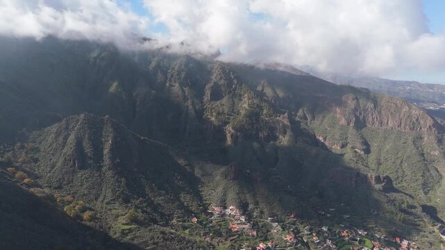 Aerial drone shot panning left across Roque Carnero mountain on Gran Canaria, Canary Islands. Capturing the rugged, lush landscape, a remote village, and terraced slopes under cloudy skies.