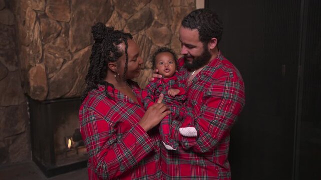 African American family Christmas time: smiling mother touches the hand of a baby held by bearded father. Matching red plaid pajamas, stone wall and lit fireplace in a cozy home at night.