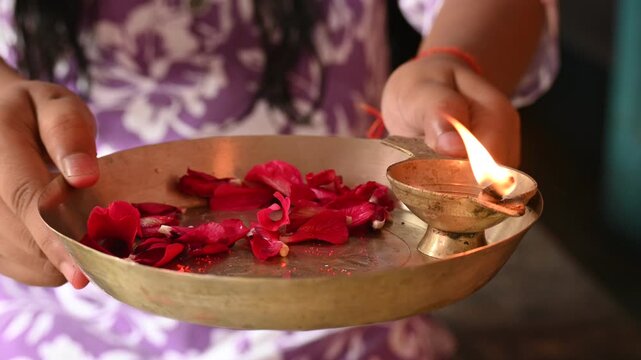 Sacred brass puja thali with lit diya and rose petals. A baby girl holds a brass puja thali featuring a glowing brass diya oil lamp and scattered fresh red rose petals. Symbolizing worship, devotion.