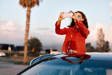 Fototapeta premium Young woman taking photo with smartphone standing through car sunroof. Sunset light, travel and modern lifestyle concept, palm tree background.