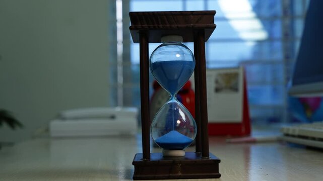 Blue sand hourglass on office desk, Blue sand hourglass on a desk in a softly blurred office or home workspace, with books, keyboard and window light in the background.