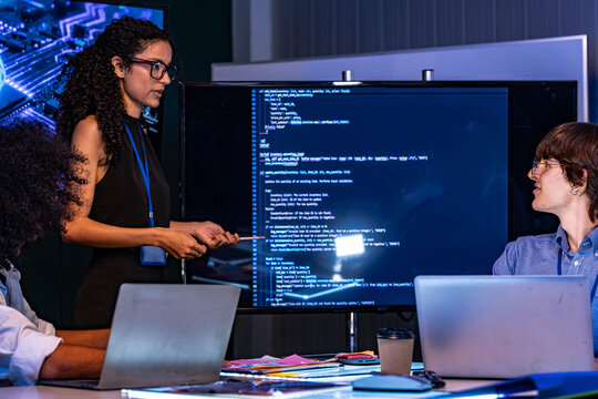 Candid moment as a curly-haired developer and a bespectacled coder review source code on a wall monitor in a modern technology office bathed in blue light, reflecting diversity and