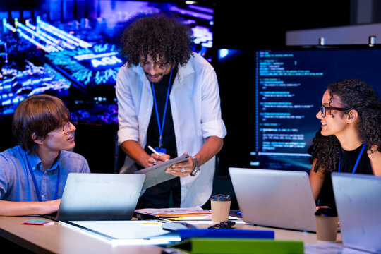 Candid moment as a diverse, multiethnic software team review work as a group at the table in a modern technology office bathed in blue light, capturing collaboration on an (frame 141)