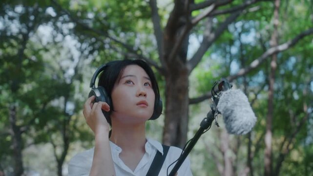 Focused Asian Woman in Headphones Holding Boom Microphone to Record Nature Sounds in Park