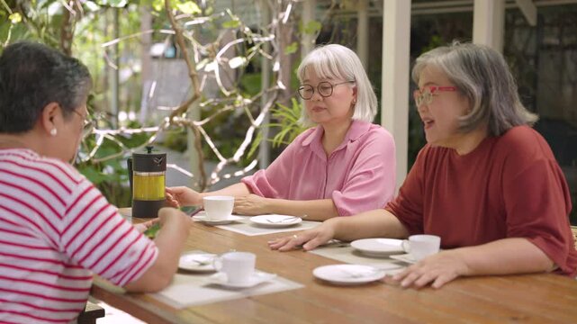 Senior friends exchange cheerful conversation beside white cups at garden cafe, sharing happiness, positive emotion, companionship through healthy aging lifestyle in leafy surroundings.