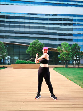 Portrait of a smiling Asian woman in black activewear and a pink cap stretching her arms before a workout in a modern urban city plaza