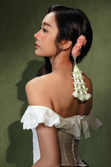 Side profile portrait of a beautiful young Asian woman wearing Malai an elegant Thai flower garland in her hair on a dark green background. © RedcupStudio