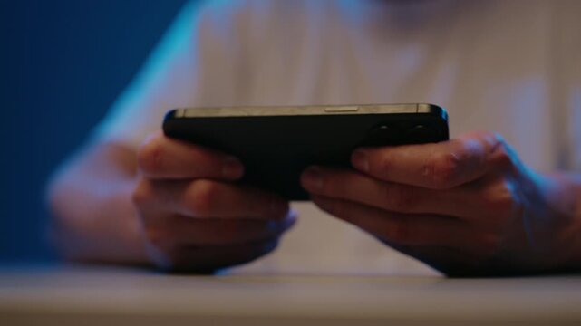 Focused man using cellphone for gaming, with hands holding device in landscape orientation sitting at table in dark room with blue and orange lighting on him, close-up, slow motion.