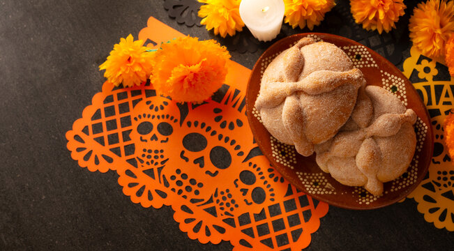 Horizontal banner of traditional Mexican Pan de Muerto sweet bread in a clay plate on a Day of the Dead offering with orange papel picado, marigold flowers, and a burning candle.