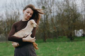 Naklejka premium A woman in a brown long sleeve top gently hugs a goat kid in an open field, wind in her hair, smiling softly. A warm, candid lifestyle moment of bonding, care, and rural nature.