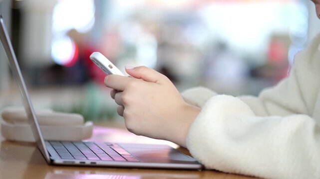 Person in cozy white sweater uses smartphone while laptop sits nearby. Focus on hands and device. Background softly blurred. Warm indoor lighting. Modern workspace scene