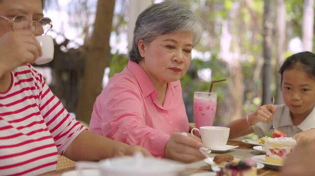 Elderly woman extends white cup across table while grandmother sits beside granddaughter, enjoying friendship, togetherness, warm companionship through active senior lifestyle outdoors.