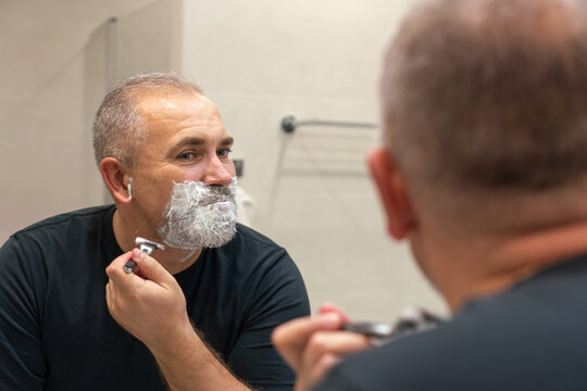 Good-looking mature man with grey hair trying to cut off his beard himself in a bathroom