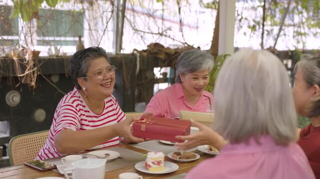 Senior woman offers red gift box across cafe table while friends share cheerful celebration while sharing warm companionship, friendship, togetherness through senior lifestyle outdoors.