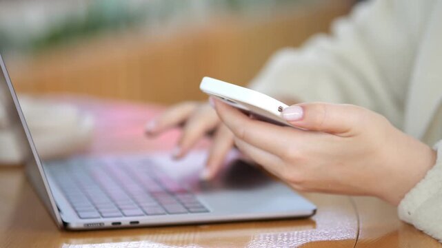 Person uses laptop and smartphone at wooden table. Hands hold white phone and type on laptop. Soft lighting with blurred background. Close-up of hands working with digital devices