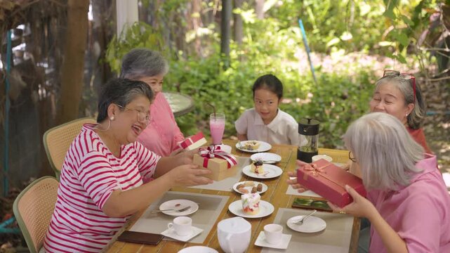 Elderly friends pass wrapped gift across table while granddaughter joins outdoor cafe celebration while enjoying friendship, social life, relaxation through active retirement lifestyle.
