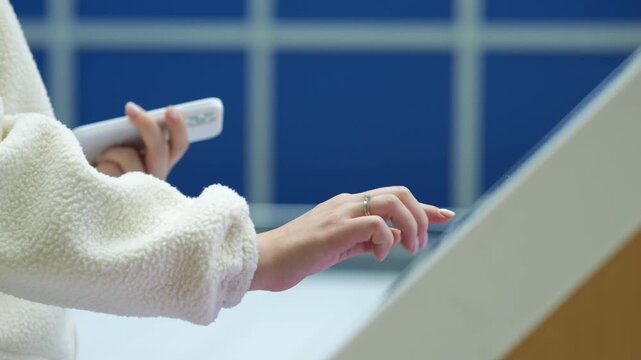 Person in white fluffy jacket holds remote. Hand gestures toward glass surface. Blue tiled wall visible behind. Modern interior with clean lines. Soft focus on hands and interaction.