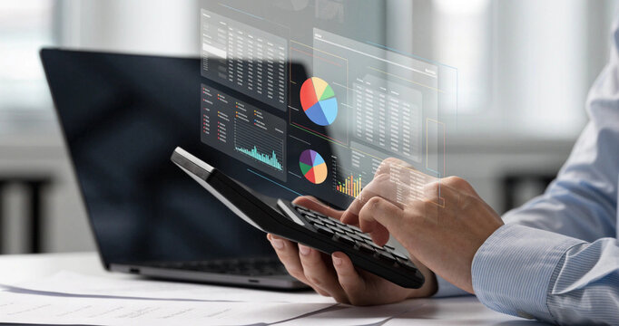 Accountant using calculator with virtual financial dashboard pie charts for tax calculation work. Financial expert using electronic calculator with floating analytics screen on office computer desk