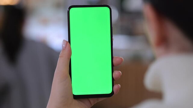 Hand holds smartphone with green screen. Background shows blurred people in indoor setting. Three keyframes show same scene slightly different angles. Person wears white shirt