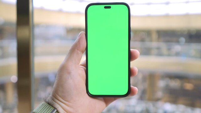 Hand holds smartphone with green screen. Background blurred mall interior visible. Person appears focused, using device. Ideal for app promotion or digital content