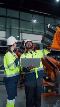 Two engineers stand beside an industrial robot arm. They wear high-visibility jackets and safety helmets. One holds a laptop. Focused and engaged