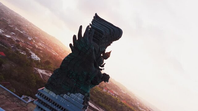 Garuda Wisnu Kencana statue in Bali standing high over the surrounding tropical landscape and distant city under a sunlit pale sky in scenic aerial drone footage