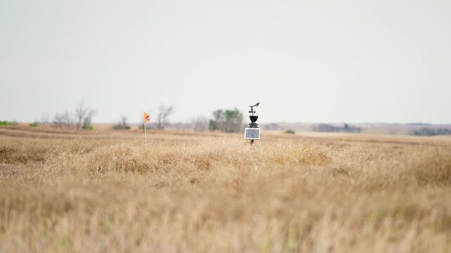 Static telephoto shot of a portable weather station with an orange flag spinning in the wind amid a golden brown harvested crop field in a rural agricultural setting