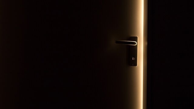 Dark room with a door ajar, a thin warm strip of light along its edge illuminating a metal lever handle and keyhole plate, copy space on the left